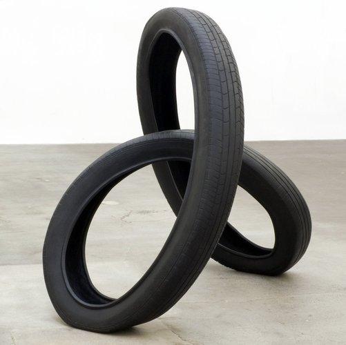 A tire, looped and twisted in a shape resembling a three-dimensional figure-eight, sits on a concrete surface. Each loop intersects with the other, forming a three-pointed structure.