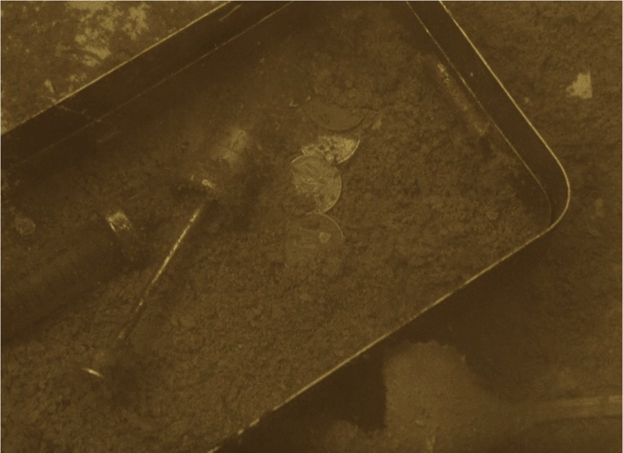 Photo of a metal box with a medical syringe and coins inside it. The box is submerged in water and partially covered in silt