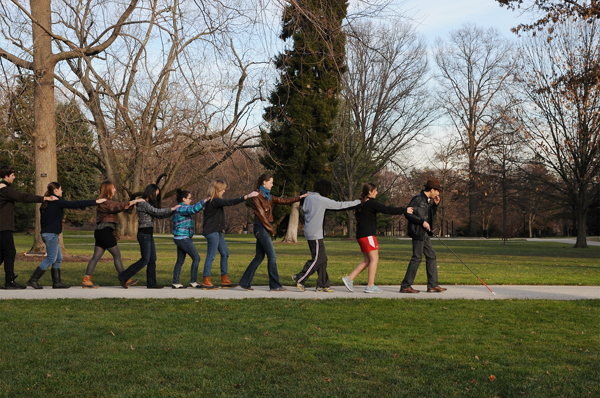 Figure 1 shows Carmen, with his signature brown derby, black leather jacket, and white-tipped cane, leading a line of students along a sidewalk bounded by grass on both sides. One of the nine students is wearing red and white gym shorts and another sports a turquoise plaid jacket. Each student is holding onto the shoulder of the person in front of her, and they appear to be walking at a smooth, rapid pace. In the background are several leafless trees and a large pine tree set against a pale blue winter sky.