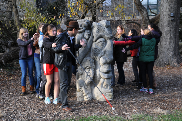 Figure 3 </strong>shows Carmen in the foreground, his right hand holding his black, red, and white-tipped cane at a sharp angle, his left hand, fingers spread, touching a rough stone sculpture about 6 feet tall and 3 feet in diameter. Carved into the stone are faces of various sizes and shapes, some human and some monstrous; the stone is partly in sunlight and partly in shadow. The students are circling the sculpture, left arms outstretched to touch it. Tree trunks and leaves are visible in the background, and the ground is covered by mulch.