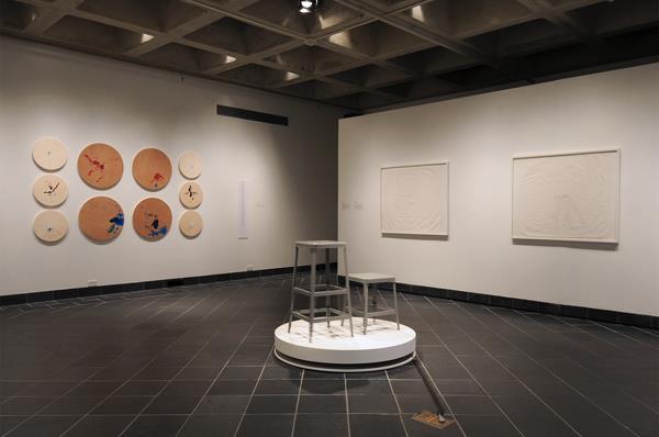 Figure 5 is a second installation shot. In the center of this shot, Laura Swanson's Display: Stools appear once again, although one might discern the movement of the stools on her rotating platform better in this photograph, as the stool legs look slightly blurred.  To the left of the stools are Christine Sun Kim's Speaker drawings #1-10 hung on the wall, composed of round wooden boards of two sizes, which contain colorful paint drip markings from her performance at the opening reception. To the right of the stools are three large drawings hung on the wall, also by Christine Sun Kim. Entitled Etudes #1-3, these are notational elements drawn with black ink onto large white sheets of paper. In this installation shot, it is hard to see any detail on the drawings.
