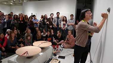 Figures 6-9</strong> show Christine Sun Kim in various stages of her performance at the opening reception for <em>What Can a Body Do?</em> at Cantor Fitzgerald Gallery. She is set up with several pieces of audio equipment, subwoofers, paint, paint brushes, other sound objects like a metronome and microphone, and many people, young and old, surround her and these objects as she engages with them. Christine wears black pants, a beige sweater and a green scarf. Christine stands as she pounds her fist on the wall in order to generate vibrations into her sound equipment.
