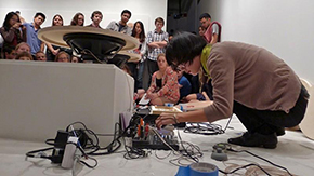 Christine is kneeling over her sound equipment placed on the ground as she plays with various cords, wires and controls.