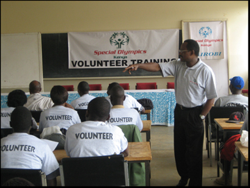 photograph of a classroom viewed from the back of the room. the chalkboard is covered by a banner reading 'special olympics kenya volunteer training'.  there is one man standing in an aisle between rows of desks where people are sitting.  the people are wearing t-shirts labeled 'volunteer'.