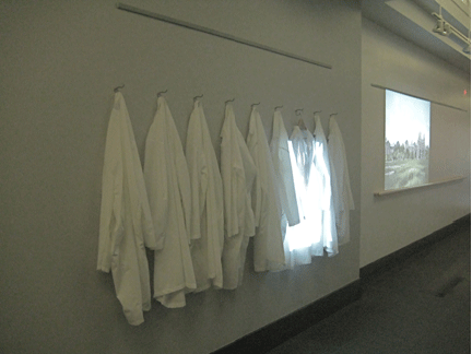 View of Still Life, from the installation artwork Admission, 2008, by J. Eisenhauer. The work juxtaposes lab coats with a digital projection of belts that bear the implication of involuntary restraint practices in psychiatric hospitals and the disabling experience of mental illness. Used with permission of Jennifer Eisenhauer and Disability Studies Quarterly