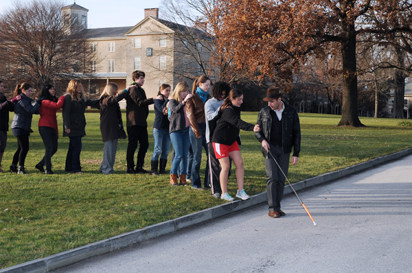 Figure 2 shows Carmen leading the same group of students from a large grassy area over a curb and onto a road. The student directly behind Carmen is feeling with her foot to find the curb and looking a bit tentative. The students behind her have stopped to wait as she navigates the curb. In the background is Founders Hall, a large fieldstone building lit by late afternoon sun, and two large trees, one already leafless and one with golden brown leaves.