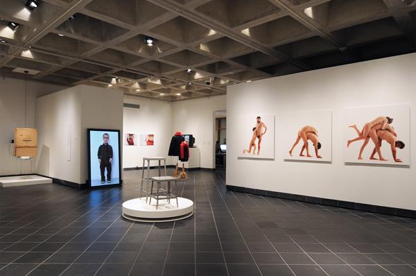 Figure 4 is an installation shot from the What Can a Body Do? exhibition at Haverford College, PA. The shot shows a variety of works ranging from sculpture to photographs and video installations. The two-dimensional images are hung on the white walls of the gallery space, while the three-dimensional sculptures sit on white pedestals throughout the space. From left to right, first, Alison O'Daniel's God's Eye, composed of a cardboard box suspended from the ceiling; Corban Walker's TV Man, a larger-than-life flat-screen television displaying a full-size portrait of Walker's four-foot body; Laura Swanson's <em>Display: Stools — grey stools sitting on a rotating circular white platform in the foreground and her Display: Clothes (dark coats and red scarves placed on free-standing dress forms) behind these; in the far background, several photographs by Chun-Shan (Sandie) Yi on the wall to the left of the dress forms; and finally Artur Zmijewski's three photographs are prominent along the wall to the right of the installation shot. These photos show nude men and women standing against a white background. One man is an amputee, and he enters into various bodily compositions with the other bodies in order to see and think about his body in alternative ways in contrast to the others.