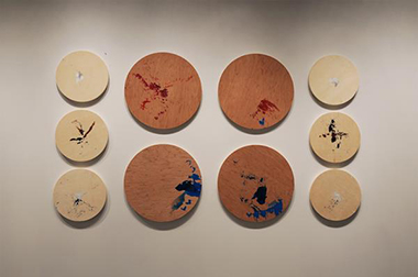 Figure 10 shows all ten of Christine's <em>Speaker Drawings</em> hung on the white wall. They are all round-shaped wooden boards, although the six smaller boards are a different type and color of wood than the four larger boards. The surface of the boards reveal scattered, abstract smears of colorful paint drips across their surfaces, creating random patterns and designs. Prominent ink colors include white, grey, red, blue and black. The boards are lined up so that the six small boards form vertical rows of three boards each, framing the left and right sides of the four large boards in the center. The large boards form two rows of two boards each, so that the overall arrangement is equally spaced and looks like one large rectangular shape hanging on the wall.