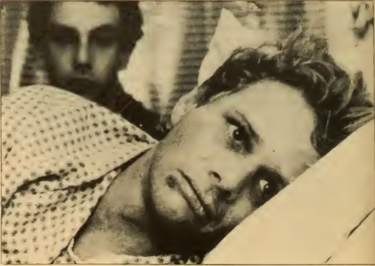 A photograph from the chest up of a man laying on his side in a bed with his eyes looking away. Another man is out of focus in the background. More description below.