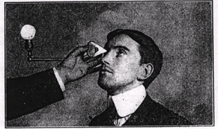 image of an early twentieth-century opthamological procedure in which a young man wearing a stiff-collared shirt, suit and, tie cranes his head slightly backward while the examiner's arm -- the only part of the examiner in the frame -- holds a conical object against the young man's eyeball