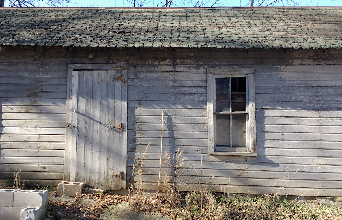 A photo of a plain wood building with a single door and window. More description below.