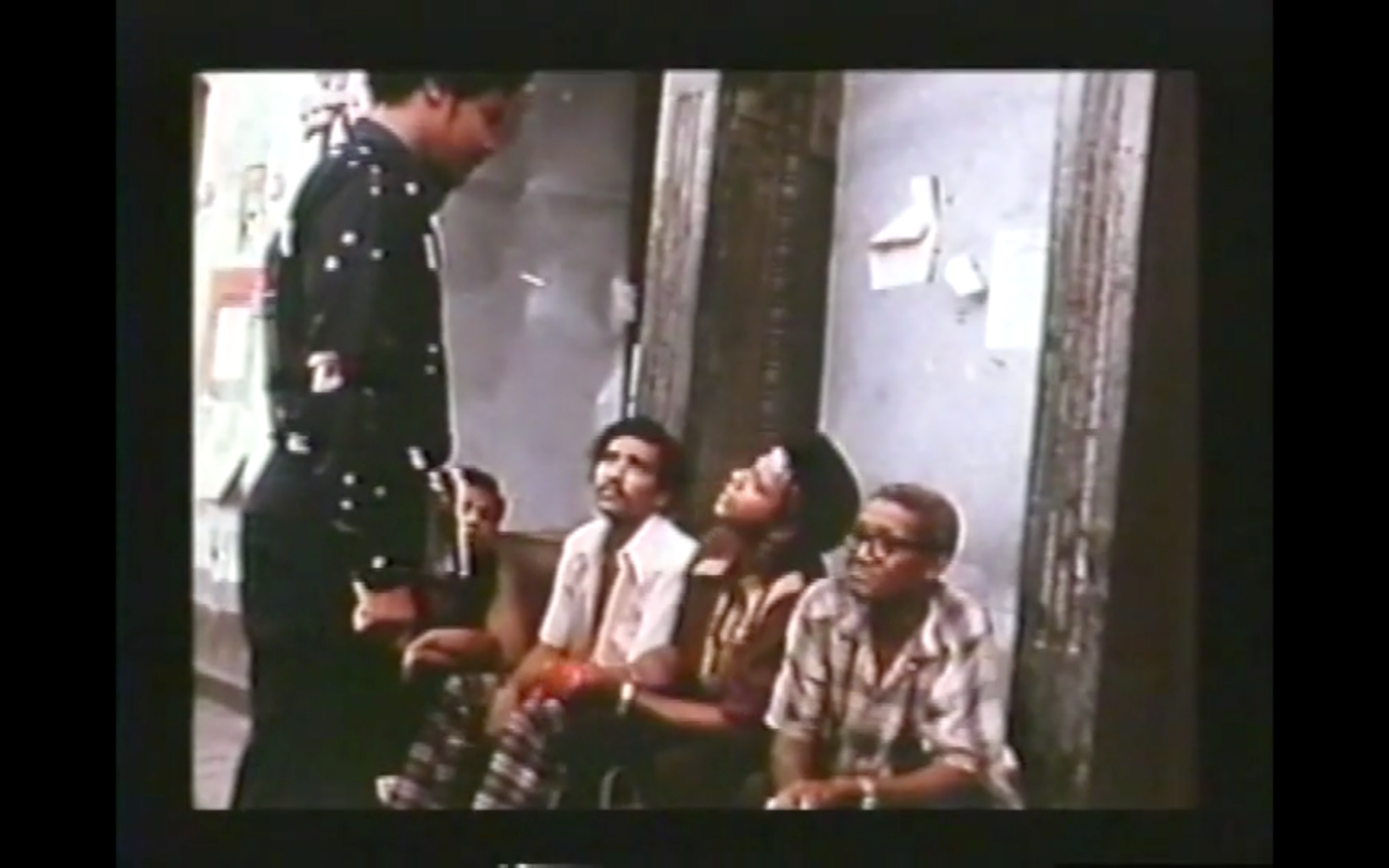 A still image from <em>Sam, Sam in Harlem</em> that shows four Black men sitting in a doorway stoop, one of whom has hailed Gaynor and Walton to come talk to them. The men wear everyday clothes; one wears a bandana with his natural style; one wears glasses and appears to be a bit older than the other three.